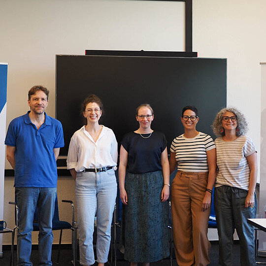 The five speakers standing in a row, smiling at the camera at the front of the room. A large TV is behind them, as well as two posters advertising the event: GIGA and MECAM.